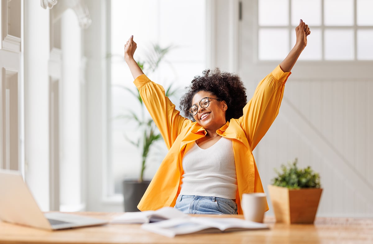 Joyful business woman freelancer smiling and rejoices in victory while sitting at desk and working at laptop screen after finishing project in home office
