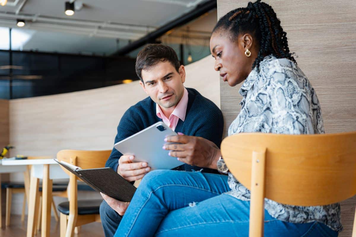 Two colleagues, a man and a woman, meeting and reviewing a digital document and notebook together