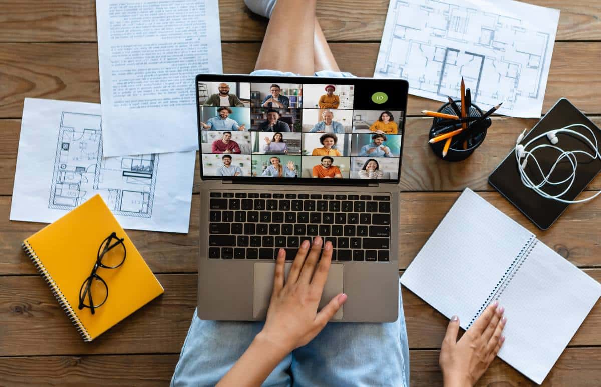 woman is sitting on a wooden floor, using a laptop to attend a conference meeting