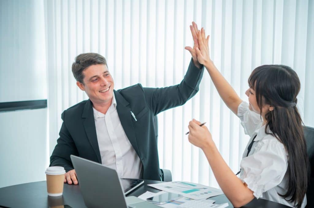 happy interracial businessperson giving high five while working near laptop at workplace