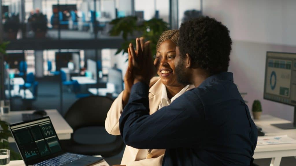 Coworkers doing high five in office after looking over positive revenue figures on PC